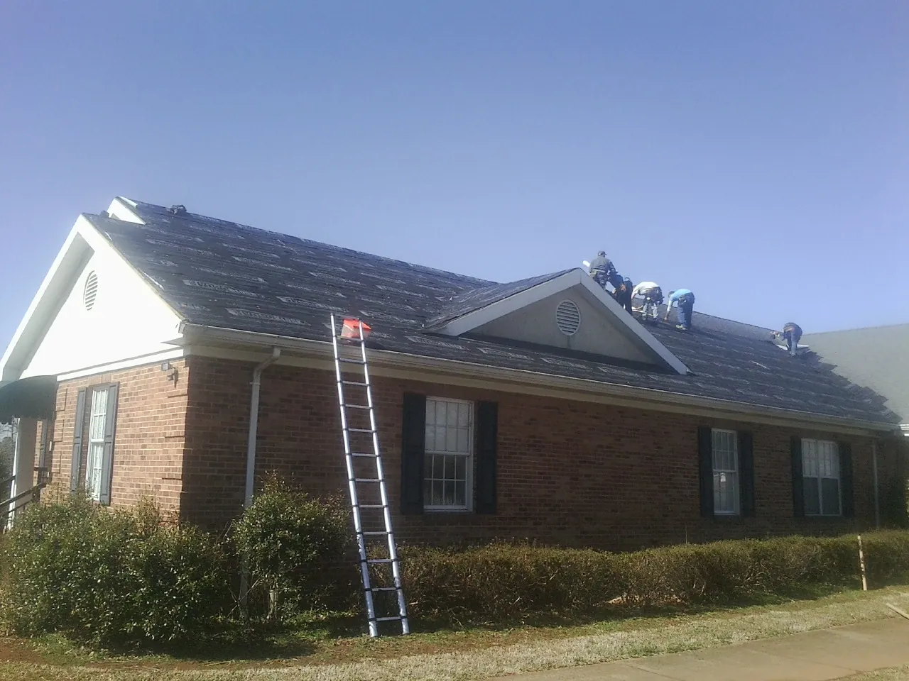Crew tearing shingles off a brick ranch roof with a ladder set — Emergency Roof Repair work by our crew in Twin Lake