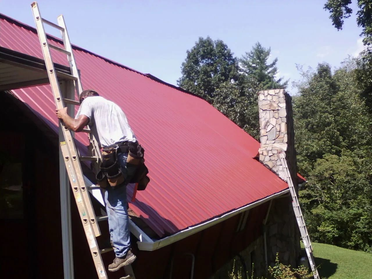 Roofer on a ladder beside a red metal roof and stone chimney — Soffit & Fascia Repair by Roofing Brothers in Twin Lake